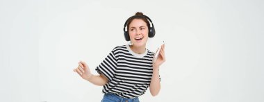 Kablosuz kulaklıkla dans eden, gözde şarkıları dinlemek için kulaklık ve cep telefonu uygulaması kullanan, beyaz arka planda poz veren şık genç bir kadının portresi..