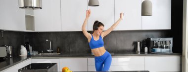 Tasasız sporcu kadın portresi, mutfakta akıllı telefonla dans etmek, kulaklıkla müzik dinlemek, antrenmandan sonra enerji dolu ve heyecanlı hissetmek..