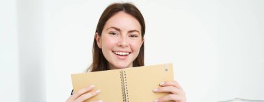 Gülümseyen, kitabını gösteren mutlu bir kadının portresi, günlük planlamacı, elinde defter, seyahatini planlayan düzenleyici notlar, beyaz arka planda izole edilmiş..
