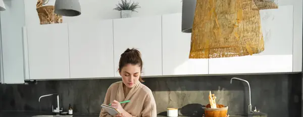 Vertical shot of young woman writing down food ideas, creating new meal, writing down grocery list or recipe in the notebook, standing in the kitchen near chopping board with fresh vegetables.