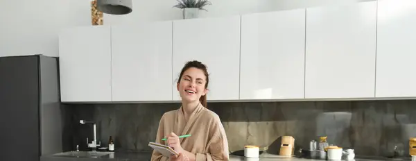 Vertical shot of young woman writing down food ideas, creating new meal, writing down grocery list or recipe in the notebook, standing in the kitchen near chopping board with fresh vegetables.