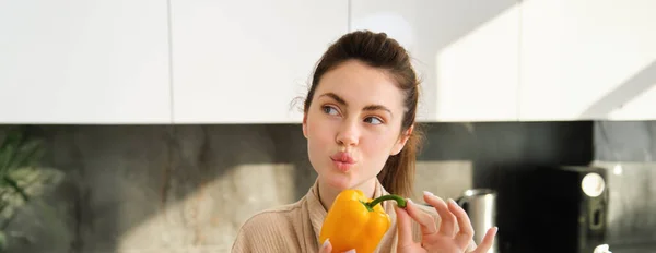 Close up of brunette woman in robe, holding yellow pepper, thinking what to cook, preparing salad or soup, making vegetarian meal with vegetables, standing in kitchen.