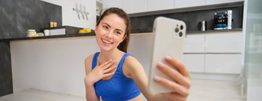 Gülümseyen güzel bir kız evde spor yaparken video kaydeder, akıllı telefona bakar, cep telefonunda selfie çeker, mavi tayt ve spor sutyeniyle evde egzersiz yapar..
