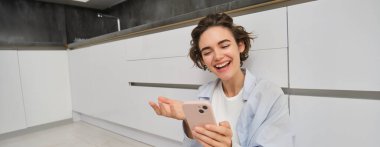 Mutlu genç bir kadın cep telefonuyla konuşur, akıllı telefon uygulamasında görüntülü sohbetler yapar, evde yerde oturur. Boşluğu kopyala