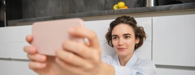 Samimi genç bir kadının portresi, akıllı telefondan selfie çekiyor, filtreli cep telefonu uygulaması kullanıyor, evde mutfak zemininde oturuyor..