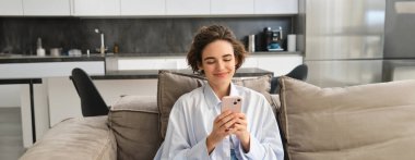 Mutlu kadın görüntüsü akıllı telefonuyla koltukta oturuyor, cep telefonu ekranına mutlu bir gülümsemeyle bakıyor, uygulamayla sohbet ediyor, internetten videolar izliyor..