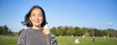 Genç kadının dikey görüntüsü ukulelesini gösteriyor, akıllı telefonu elinde tutuyor ve kameraya bakıyor. İnsanlar ve hobiler konsepti