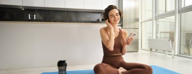Aktif giyimli spor bir kadın görüntüsü, evde yoga minderinde oturuyor, akıllı telefonu tutuyor, egzersiz uygulaması kullanıyor, çevrimiçi egzersiz arıyor. Boşluğu kopyala