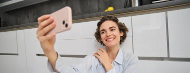 Genç bir kadının portresi mutfak zemininde telefonuyla oturuyor, akıllı telefonuyla fotoğraf çekiyor uygulama filtreleriyle, cep telefonuyla poz veriyor..