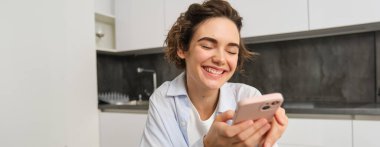 Cep telefonu tutan ve gülümseyen modern esmer kadının portresi. Kız evde akıllı telefonuyla mutfakta oturuyor, teslimat için uygulama kullanıyor, online alışveriş yapıyor..