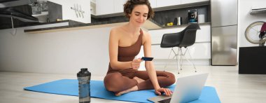 Genç fitness kızı, evde bayan sporcu, mutfak zemininde antrenman, dizüstü bilgisayarla kredi kartı tutma, online eğitim oturumu satın alma, ev antrenmanı için beden eğitimi başvurusu.