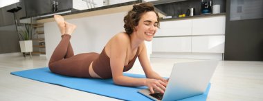 Kadın sporcu portresi, kız antrenmanı, dizüstü bilgisayarda spor videoları, lastik paspasın üzerine uzanmak ve kapalı yoga seansı yapmak. Spor ve refah kavramı