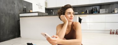 Cep telefonlu sportif bir kızın portresi, yoga minderinde oturmak, antreman sırasında akıllı telefon kullanmak, telefonda spor salonunu izlemek. Spor ve refah kavramı