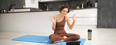 Gülümseyen genç fitness kadını, eğitmen tablette el sallıyor, evdeki sporla ilgili video blogunu kaydediyor, çalışma sınıfına merhaba diyor, esneme hareketleri yapıyor ve spor egzersizleri yapıyor..