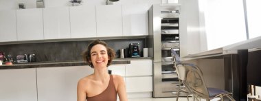 Dikey çekimde, genç bir kadın evinde oturmuş yoga yapıyor, lotus pozu veriyor ve meditasyon yapıyor, rahatlamış ve mutlu, gülümsüyor..