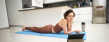 Gülümseyen genç sporcu kadın, yoga minderinde uzanan, tablete bakan, spor eğitimi alan, yoga dersi alan, kablosuz kulaklık takan, mutfakta esneyen ve egzersiz yapan..