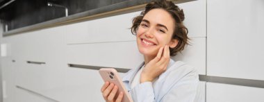 Evinde telefonu olan mutlu bir kadının portresini yakın çekim, yüzüne dokunuyor ve hafifçe gülümsüyor, mobil uygulama kullanıyor. Teknoloji ve insanlar kavramı.
