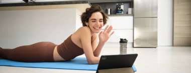 Spor ve yaşam tarzı. Genç fitness kadını, beden eğitimi hocası tablette el sallıyor, çevrimiçi video sınıfına katılıyor, yoga ve farkındalık egzersizi yapıyor, evde antrenman yapıyor..