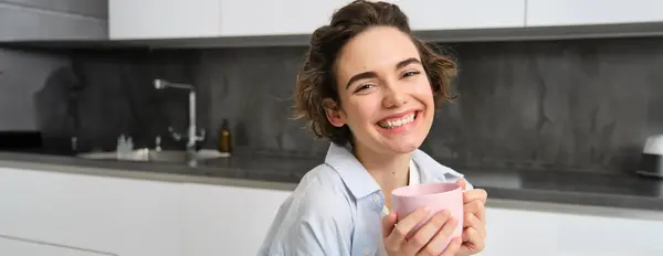 stock image Happy brunette woman drinking coffee at home. Girl with pink cup, sits in her kitchen and enjoys relaxing morning weekend.
