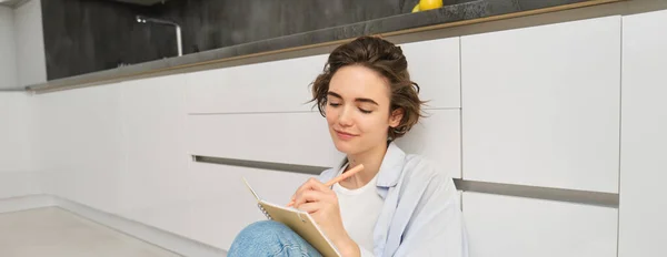 Portrait of beautiful young woman writing in journal, adding notes in planner, sitting on floor and thinking, reads her diary, smiles with pleased face expression.