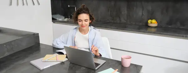 Image of young productive woman works from home, does distance learning, learns online course on her laptop in kitchen, writes down information, watches webinar, attends work meeting remotely.