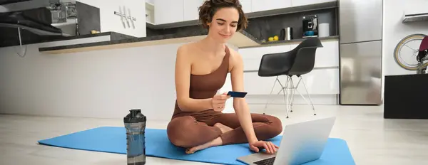 Image of young fitness girl, female athlete at home, workout on kitchen floor, holding credit card with laptop, purchasing online training session, gym application for home workout.