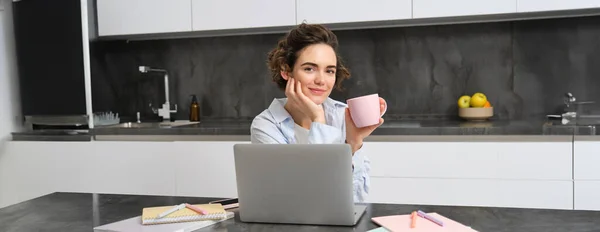 Self-employed woman works from homes, sits with laptop and drinks coffee in kitchen, surrounded with documents. Girl student studies remote, does project on computer, holds a cup.