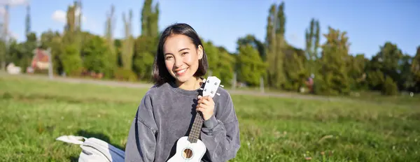 Smiling asian girl learns how to play ukulele on laptop, video chat with music teacher, sitting with instrument in park on grass.