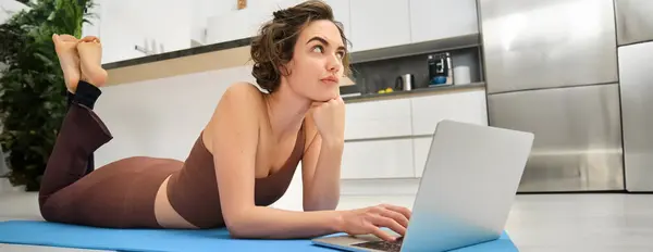 Fitness girl lying on rubber mat with thoughtful face, using laptop, fitness instructor thinking, planning online class, workout at home in kitchen. Sport and recreation concept