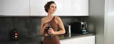 Spor ve sağlıklı yaşam tarzı. Gülümseyen spor elbiseli bir kadının portresi, mutfak tezgahının yanında duruyor, su şişesinden içiyor, spor salonuna hazırlanıyor..
