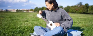 Çimenlerde ukulele çalan Asyalı kız şarkı söylüyor, parkta battaniyeye oturuyor, güneşli bir günde dışarıda dinleniyor..