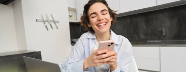 Evde akıllı telefonunu kullanan genç bir kadının görüntüsü. Kız cep telefonuyla mutfakta oturuyor ve gülümsüyor..