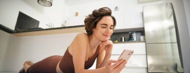 Spor kızı imajı yoga paspasında dinleniyor, evde egzersiz yapıyor, akıllı telefonunu kullanıyor, online fitness dersleri arıyor, eğitim için mobil uygulama arıyor..