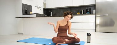 Kadın, jimnastik hocası lastik mindere oturuyor, yoga kanepesi çevrimiçi grupla konuşuyor, tablet üzerinde kayıt dersi, spor salonu dışında uzaktan eğitim, evden antrenman..