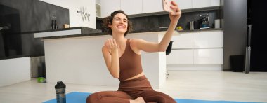 Yaşam tarzı ve egzersiz. Modern fitness kızı yoga minderinde selfie çekiyor, egzersizlerini evde yapıyor ve akıllı telefondan fotoğraf çekiyor, canlı video, telefonda video kaydediyor..