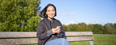 Teknoloji ve insanlar. Akıllı telefon kullanan genç bir kadın, parkta bankta oturuyor, akıllı telefona bakıyor, mesaj gönderiyor ya da ağ kuruyor..