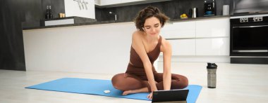 Gülümseyen genç bir kadının portresi, kauçuk yoga minderinde fitness eğitmeni, tabletten online eğitim sınıfına katılıyor, kablosuz kulaklıkla video dinliyor, evde yerde egzersiz yapıyor..