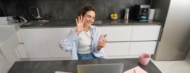 Dost canlısı genç bir kadın, dizüstü bilgisayara el sallıyor, video sohbetleri yapıyor, akıllı telefonu tutuyor, internette sohbet ediyor..