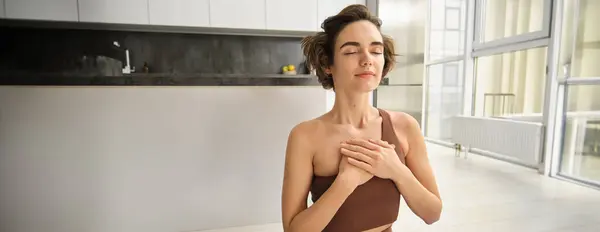 stock image Sukhasana Hands To Heart. Portrait of young brunette fitness woman, girl holds hans on chest, doing yoga at home in sport leggins and bra, sitting on floor indoors.