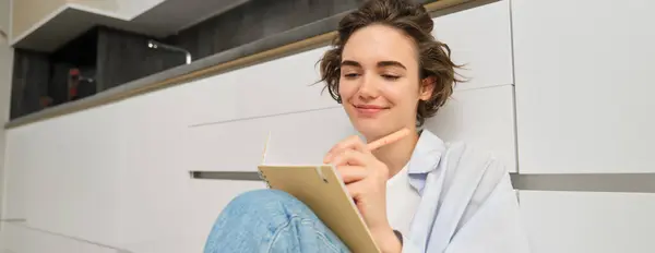 Hobbies and lifestyle concept. Happy young woman sits on floor with pen and notebook, wirtes in her diary, makes notes in her journal, plans her schedule, draws on paper.