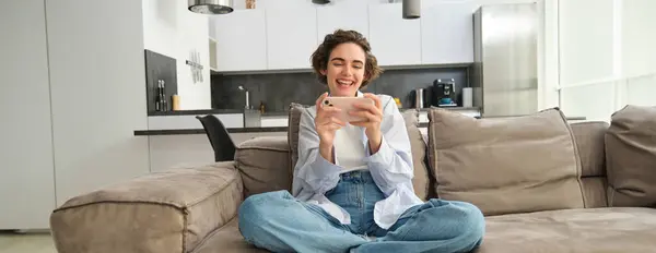 Portrait of young girl gamer, plays mobile video game on smartphone, sits on sofa with excited face expression.