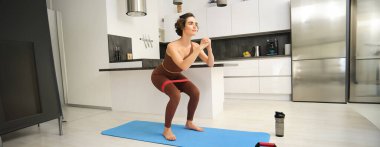 Evde fitness var. Genç sporcu kadın, aktif giyimli kız, mutfakta çömelme hareketleri yapıyor, lastik paspasın üzerinde egzersiz yapıyor, lastik bant kullanarak egzersiz yapıyor..