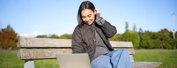 Young brunette girl sits in park with laptop, watches video or browses internet, rests outdoors on bench in sunny park.