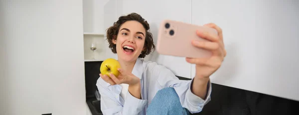 Beautiful woman eating an apple and recording herself on video. Cute girl with fruit, posing for photo, taking selfie in kitchen in happy mood.