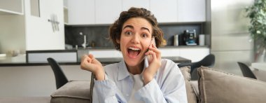 Şaşırmış esmer kadının yakın portresi, heyecandan koltuğa atlıyor, telefonlara cevap veriyor, telefondan harika haberler duyuyor, şaşırmış ve mutlu bir şekilde tepki veriyor..