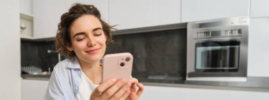 Cep telefonu tutan ve gülümseyen modern esmer kadının portresi. Kız evde akıllı telefonuyla mutfakta oturuyor, teslimat için uygulama kullanıyor, online alışveriş yapıyor..