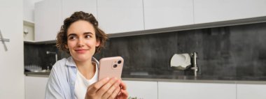 Cep telefonu tutan ve gülümseyen modern esmer kadının portresi. Kız evde akıllı telefonuyla mutfakta oturuyor, teslimat için uygulama kullanıyor, online alışveriş yapıyor..
