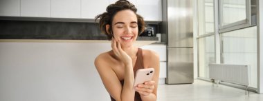 Aktif giyimli spor bir kadın görüntüsü, evde yoga minderinde oturuyor, akıllı telefonu tutuyor, egzersiz uygulaması kullanıyor, çevrimiçi egzersiz arıyor. Boşluğu kopyala