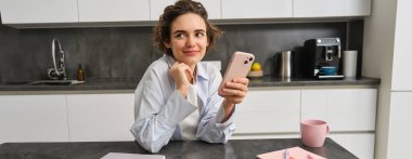 Güzel gülümseyen bir kadının portresi akıllı telefonuyla evde oturuyor, cep telefonu uygulaması kullanıyor, erzak siparişi veriyor..