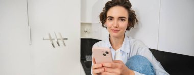 Gülümseyen esmer kız akıllı telefonuyla evde oturuyor, cep telefonu uygulamasından sipariş veriyor, online alışveriş uygulaması kullanıyor..